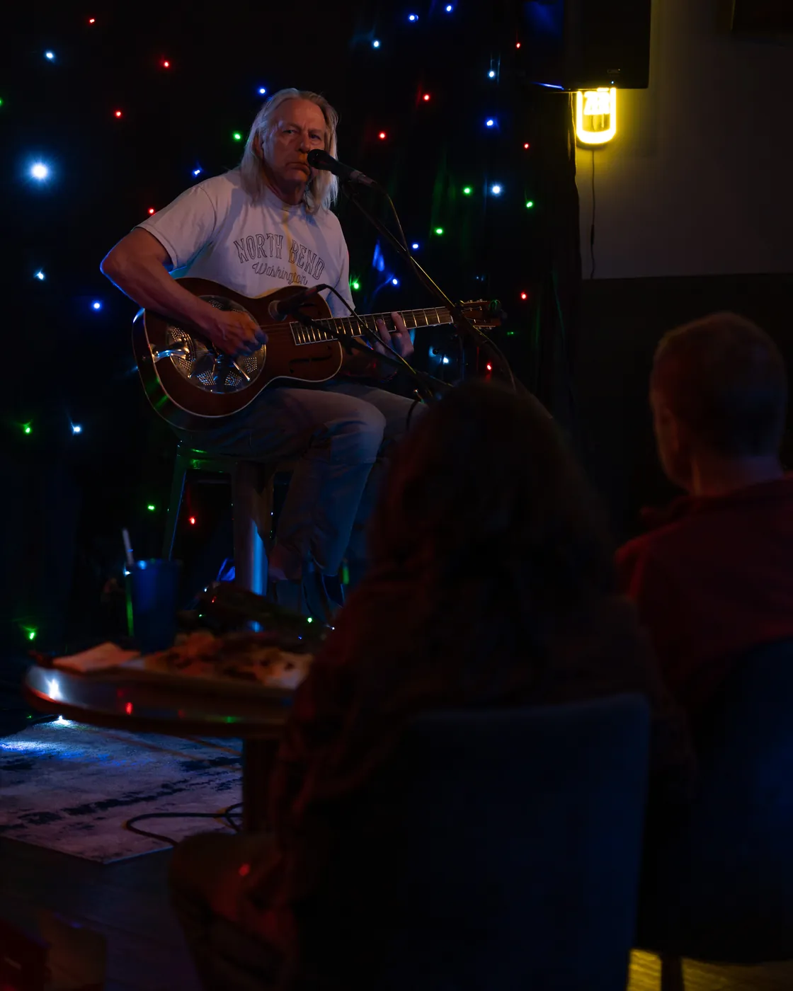 Singer-songwriter playing acoustic guitar on a small stage under colorful lights