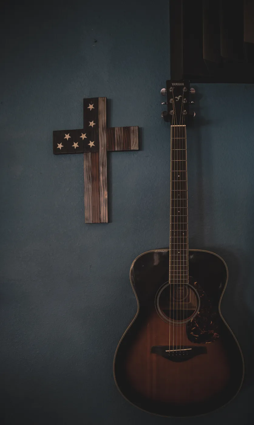 Acoustic guitar hanging beside a hand-painted American flag cross on a blue wall